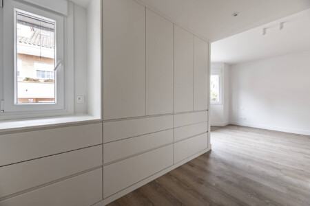 Newly installed bedroom dressing room with white wooden cabinets and drawers, light wooden floor and access to an empty room with several windows